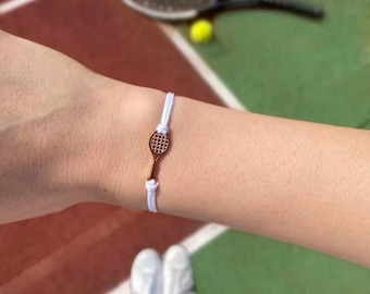 May include: A white cord bracelet with a bronze tennis racket charm. The bracelet is tied with knots and is worn on a wrist. A tennis court and tennis equipment are in the background.