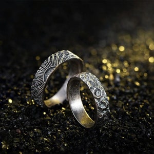 Puede incluir: Dos anillos de plata con superficies texturizadas. Un anillo presenta un diseño de sol, mientras que el otro anillo presenta un diseño de luna.