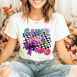 Puede incluir: Camiseta blanca con la frase "GAMER GIRL" en letras coloridas y divertidas. El diseño incluye un lazo morado, un patrón de tablero de ajedrez y dos gráficos de cerezas rosas, creando un aspecto divertido y vibrante.