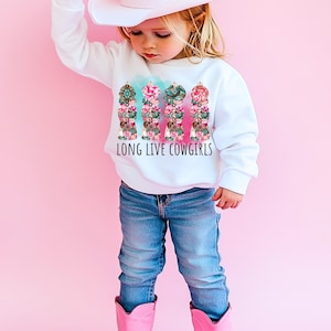 May include: A white sweatshirt with the text "LONG LIVE COWGIRLS" and a colorful graphic of four decorative columns. The columns feature floral and turquoise designs. The child is wearing blue jeans, pink boots, and a white cowboy hat.