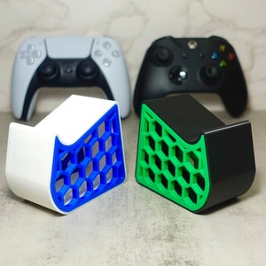 May include: Two 3D printed controller stands, one white with a blue honeycomb design and one black with a green honeycomb design. The stands are designed to hold video game controllers.