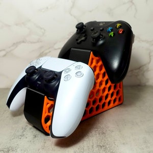 May include: A black and white video game controller sits on a black and orange charging stand. A second black video game controller is also on the stand.