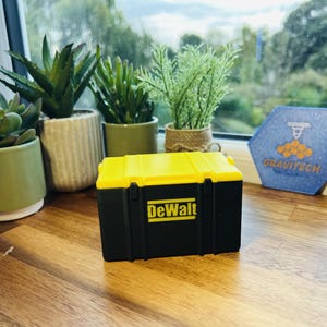 May include: A black and yellow tool box with the DeWalt logo on the front. The box is sitting on a wooden surface in front of a window.