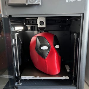 May include: A red and black Deadpool mask is inside a 3D printer. The printer is gray with the text "Bambu Lab P1S" on the side. The printer bed dimensions are 256x256x256mm.