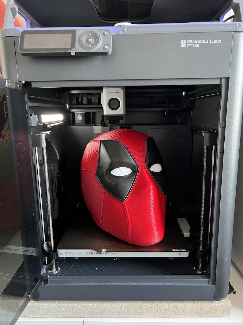 May include: A red and black Deadpool mask is inside a 3D printer. The printer is gray with the text "Bambu Lab P1S" on the side. The printer bed dimensions are 256x256x256mm.