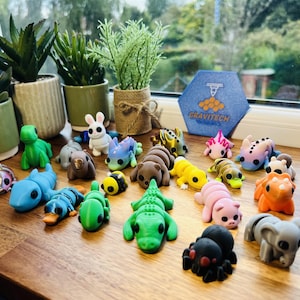 May include: A collection of colorful, small animal figurines, including a green alligator, a pink pig, and a gray elephant. The figurines are arranged on a wooden surface near potted plants and a window.