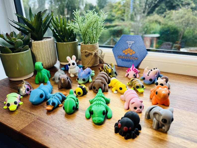 May include: A collection of colorful, small animal figurines, including a green alligator, a pink pig, and a gray elephant. The figurines are arranged on a wooden surface near potted plants and a window.