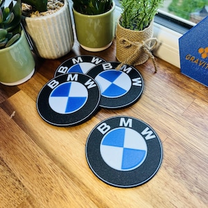 May include: Four black and blue BMW logo coasters with a white and blue checkered design. The coasters are round and have the BMW logo in white text. The coasters are on a wooden surface.