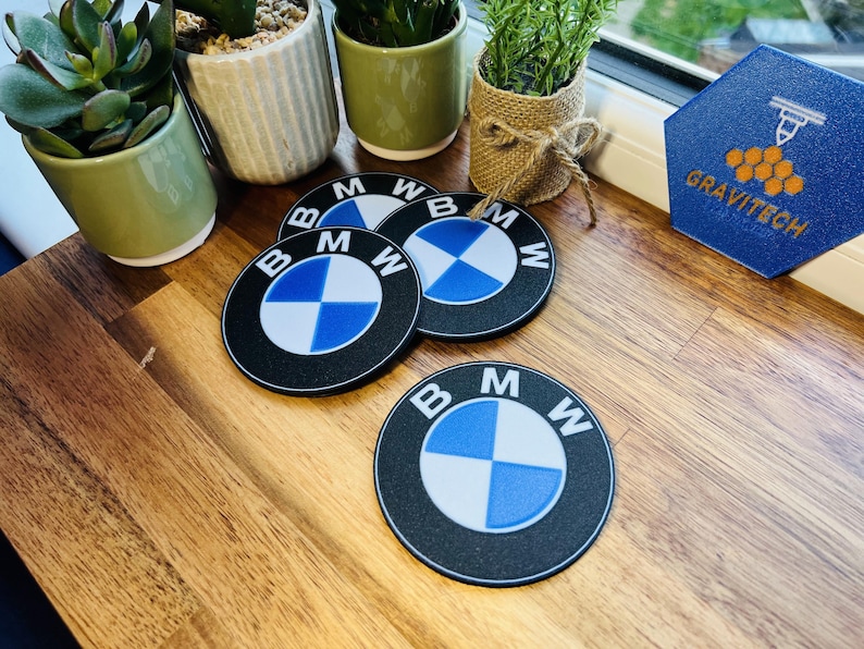 May include: Four black and blue BMW logo coasters with a white and blue checkered design. The coasters are round and have the BMW logo in white text. The coasters are on a wooden surface.