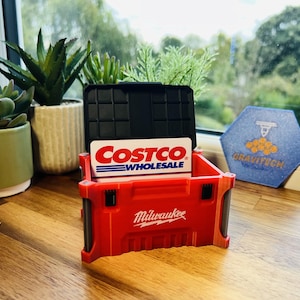 May include: A red Milwaukee-branded toolbox-style card holder with a black lid, holding a Costco Wholesale card. The holder sits on a wooden surface, with potted plants and a window in the background. A blue and white hexagon-shaped item is also visible.