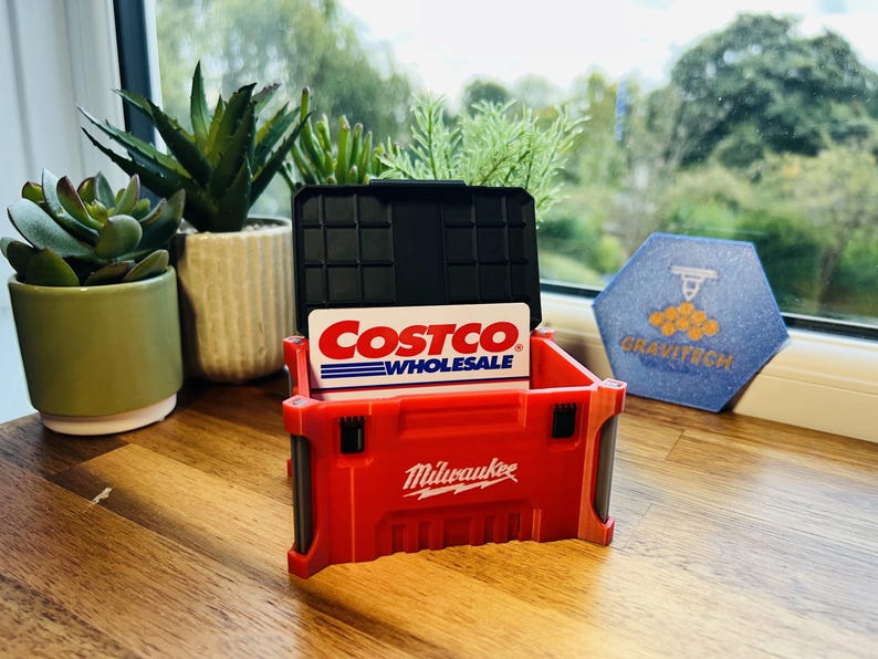 May include: A red Milwaukee-branded toolbox-style card holder with a black lid, holding a Costco Wholesale card. The holder sits on a wooden surface, with potted plants and a window in the background. A blue and white hexagon-shaped item is also visible.