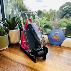 May include: Clear phone case with a black phone inside, displayed on a pink and black stand. The stand is on a wooden surface, with small potted plants and a blue hexagon-shaped logo in the background.