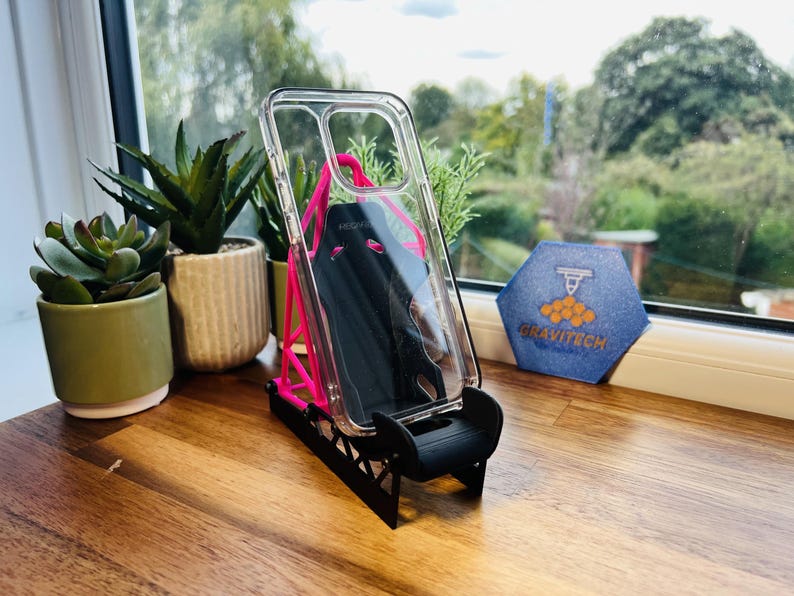 May include: Clear phone case with a black phone inside, displayed on a pink and black stand. The stand is on a wooden surface, with small potted plants and a blue hexagon-shaped logo in the background.