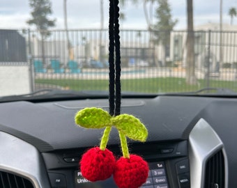 May include: A crocheted cherry car charm with green leaves and red cherries hanging from a black string. The charm is hanging from the rearview mirror of a car.