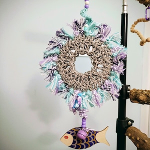 May include: A bird toy featuring a woven, circular centerpiece with hanging tassels in shades of gray, teal, and purple. A wooden fish with purple and gold details hangs below. The toy is suspended by a purple clip and beads.