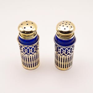 May include: A pair of salt and pepper shakers. Each shaker has a gold-colored top with small holes, a vibrant blue glass body, and a decorative gold-colored metal filigree cage. The shakers are displayed against a white background.