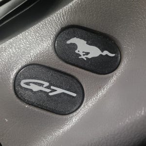 Puede incluir: Primer plano de un detalle interior de un coche negro y gris con dos botones. El botón superior tiene un logotipo de un caballo corriendo, y el botón inferior tiene el texto "GT" en blanco.