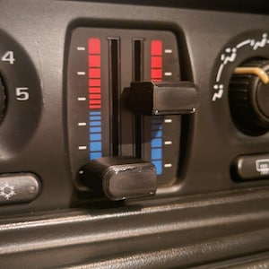 May include: Close-up of a car's dashboard with climate control features. The image shows a black panel with red and blue temperature indicators, sliders, and a rotary dial. The temperature indicators are marked with red for hot and blue for cold.
