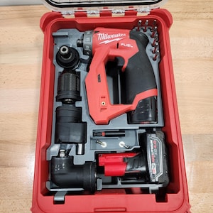 May include: A red and black Milwaukee FUEL drill kit in a red case. The kit includes a drill, battery, and various attachments. The case is open, revealing the tools neatly organized within. The drill is red and black, with the Milwaukee logo visible.