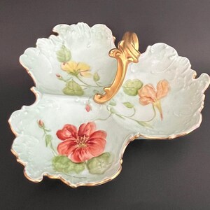 May include: A three-sectioned, light green ceramic serving dish with a gold handle. The dish is decorated with hand-painted flowers in shades of red, yellow, and orange. The edges are trimmed with gold.