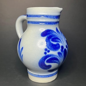 Op de afbeelding: Kleine, gebroken witte keramische kruik met een handvat en tuit. De kruik is versierd met blauwe bloemmotieven en horizontale blauwe strepen. De basis is een lichtere tint gebroken wit. De kruik is ongeveer 15 cm hoog.