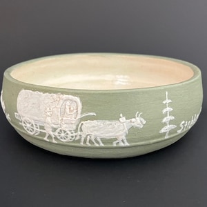 May include: A sage green ceramic bowl with a cream interior. The exterior features a white bas-relief design of a covered wagon pulled by oxen, a person, and a tree. The bowl has a rounded shape.