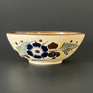 May include: A ceramic bowl with a beige exterior and a brown rim. The bowl is decorated with a floral pattern featuring blue, brown, and yellow flowers and green leaves. The bowl is likely handmade and suitable for serving food.
