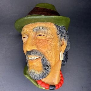 Puede incluir: Una placa de pared de cerámica con la cabeza de un hombre con un sombrero verde, un collar de cuentas negras y rojas y un pendiente blanco. El hombre tiene barba y bigote y sonríe.