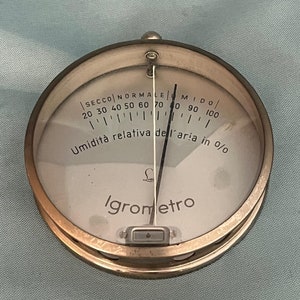 Può includere: Un igrometro rotondo di colore argento con un ago nero e un quadrante bianco. Il quadrante ha le parole "SECCO NORMALE MIDO" e "Umidità relativa dell'aria in 0/0" stampate su di esso. L'igrometro ha anche la parola "igrometro" stampata su di esso.