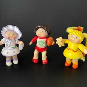 Peut inclure: Trois petites poupées en plastique avec des visages ronds et des cheveux en laine. L'une porte un bonnet et une robe blancs à cœurs violets, une autre un haut et un short de sport rouges avec un ballon de football, et une robe jaune avec un ours.