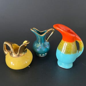May include: Three small ceramic pitchers. The left pitcher is yellow with a gold-toned rim and handle. The centre pitcher is iridescent teal with a gold-toned rim and handle. The right pitcher features a tri-colour design of orange, yellow, and blue.