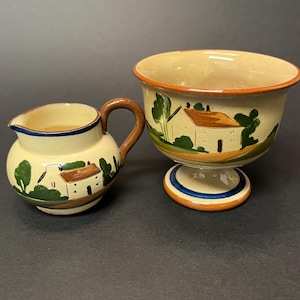 Puede incluir: Un pequeño juego de jarra de cerámica y cuenco con pedestal. Las piezas de color crema presentan un diseño pintado a mano de una casa con árboles verdes. La jarra tiene un asa marrón y ribete azul. El cuenco tiene un borde marrón y una base azul y naranja.