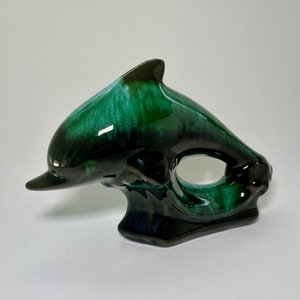 Puede incluir: Una figurita de delfín de cerámica esmaltada en verde y negro. El delfín está saltando fuera del agua y tiene un diseño de remolino blanco en su cuerpo.