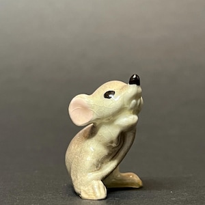May include: A small, ceramic mouse figurine in a seated pose, looking upwards. The mouse has a light beige body with a black nose and eyes, and pink ears. The figurine has a crackle glaze finish.