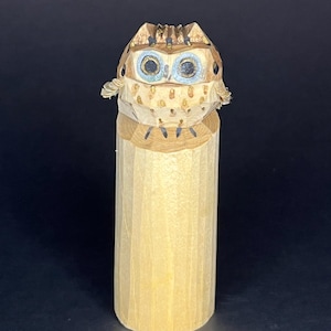 Könnte beinhalten: Eine Holzfigur eines Eulen mit blauen Augen und goldenen Akzenten. Die Eule sitzt auf einem Holzsockel.