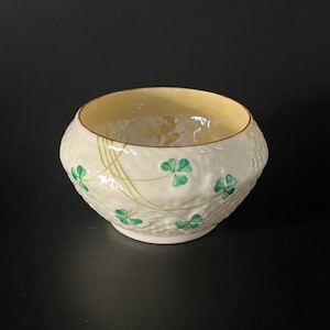 Puede incluir: Un cuenco de cerámica blanca con exterior texturizado e interior amarillo. El cuenco está decorado con tréboles verdes y enredaderas verdes. El cuenco es redondo y tiene una abertura ancha.
