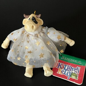 Könnte beinhalten: Eine skurrile Kuhfigur, die ein weißes Kleid mit goldenen Sternen trägt. Die Kuh hat einen schwarzen Fleck um ein Auge und braune Hörner. Ein Etikett lautet "Kathleen Kelly".