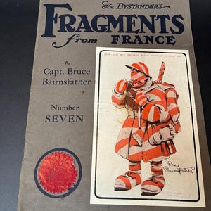 K&ouml;nnte beinhalten: Ein Vintage-Buchcover mit dem Titel "Fragments from France" von Capt. Bruce Bairnsfather. Das Cover ist grau mit dunkelblauem Text und einem roten Siegel. Eine gerahmte Illustration zeigt eine Cartoonfigur in rot-wei&szlig;en Streifen.