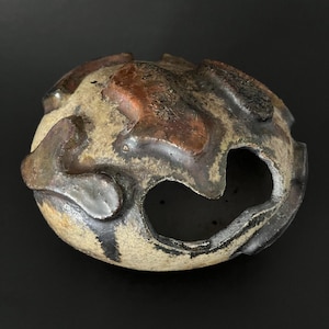 Pode incluir: Uma esfera de cerâmica artesanal com uma superfície texturizada. A cerâmica apresenta uma mistura de cores, incluindo bege, castanho e preto, com um grande recorte aberto. O design único sugere um propósito decorativo ou artístico.
