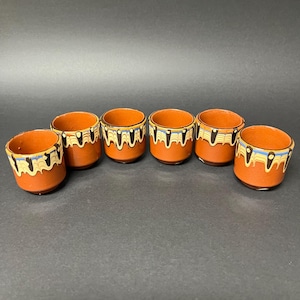 Peut inclure: Six petites tasses en céramique de couleur terre cuite. Chaque tasse présente une bande décorative avec des motifs noirs, jaunes et bleus. Les tasses sont disposées en rangée sur un fond sombre.