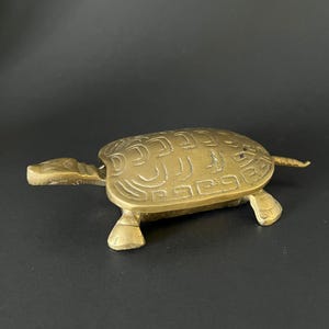 May include: A brass turtle ornament with engraved patterns. The turtle has a rounded shell, a small head, and four short legs. The figurine is a warm gold colour and sits on a dark surface. The turtle's shell has a textured design.