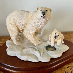 Pode incluir: Estatueta de urso polar com filhote, sobre um bloco de gelo esculpido. Os ursos são creme, com detalhes de pelo, montados numa base de madeira escura. Uma representação da vida selvagem.