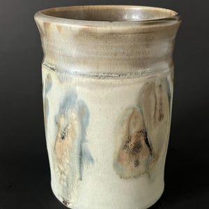 May include: A handcrafted ceramic tumbler with a distinctive, naturalistic design. The top section is a light tan, fading into a creamy white base. The surface displays abstract, flowing patterns in blue and brown, giving a rustic appearance.