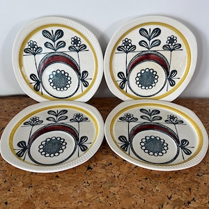 以下が含まれることがあります： クリーム色の背景に4つの楕円形のセラミックプレート。各プレートには、濃いティールサークル、赤い三日月、黒とティールの花のモチーフが中心にデザインされています。プレートには黄色の縁があります。