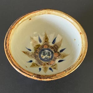 以下が含まれることがあります： 中央に手描きの太陽のモチーフが施された、クリーム色の内側の小さな丸い陶器のボウル。太陽には濃い青色の目を持つ顔が描かれており、茶色の放射状の縁取りと小さな青い涙の形をしたアクセントが特徴です。