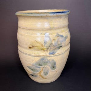 May include: A handmade ceramic vase with a beige and blue floral design. The vase has a cylindrical shape with horizontal ridges and a slightly flared rim. The floral pattern features blue and green leaves and stems. The vase is approximately 7 inches tall.