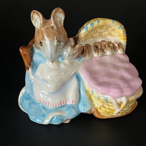 以下が含まれることがあります： 青いドレスを着たネズミの陶器製置物。ピンクの毛布が入ったバスケットを持っています。バスケットは黄色で青いアクセントが付いています。ネズミは茶色の毛皮と白いエプロンをしています。可愛らしい装飾品です。