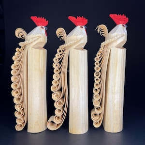 Könnte beinhalten: Drei hölzerne Hahnfiguren mit roten Kämmen und weißen Körpern. Jeder Hahn hat einen langen, gekräuselten Schwanz aus Holzspänen. Die Hähne stehen auf Holzsockeln.