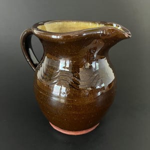 May include: A small, brown ceramic jug with a handle and spout. The jug has a glossy finish and a reddish-brown base. The interior is a lighter, yellowish-brown colour. The jug is likely handmade and suitable for serving liquids.