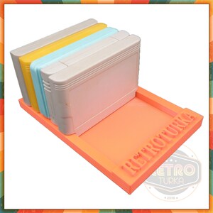 Könnte beinhalten: Ein leuchtend orangefarbener Spielkassettenhalter aus Kunststoff mit dem aufgedruckten Text "RETROTURKA". Der Halter ist mit vier Spielkassetten gefüllt, einer gelben, einer hellblauen, einer hellblauen und einer weißen.